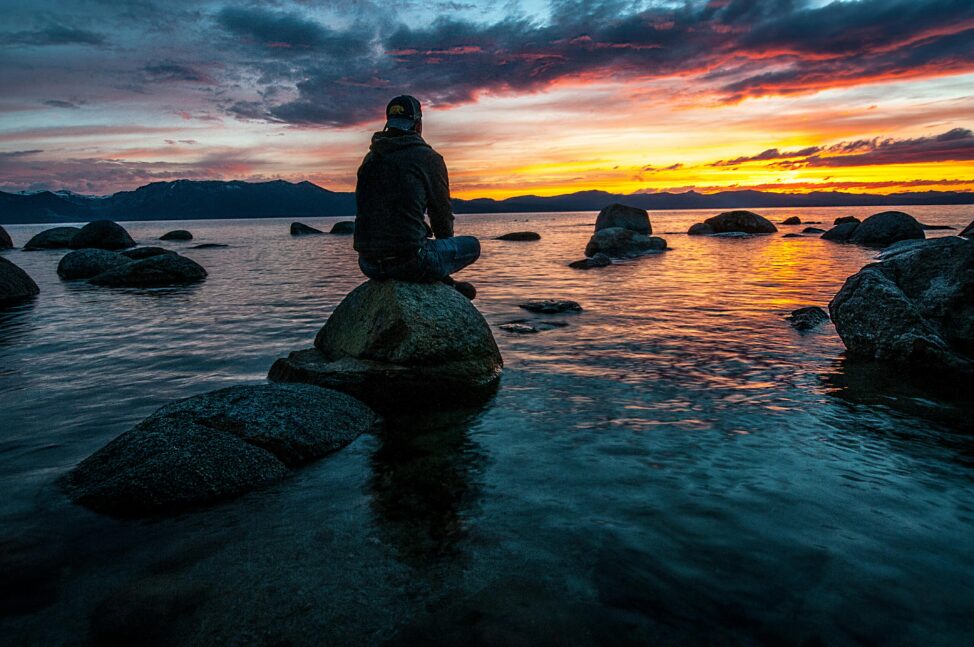 a man in a hoodie sitting cross legged on a rock in a body of water, watching a sunset - a different kind of year end review